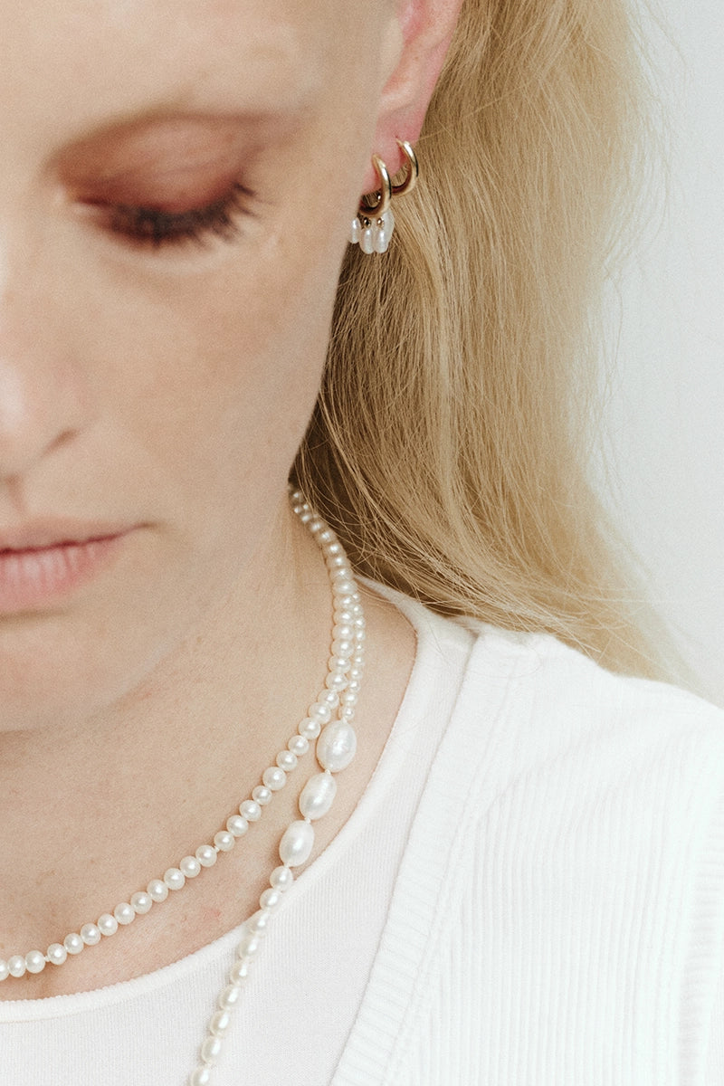 Woman wearing stacked yellow gold huggie hoop earrings with white pearl drops and layered white pearl necklaces – Daure collection